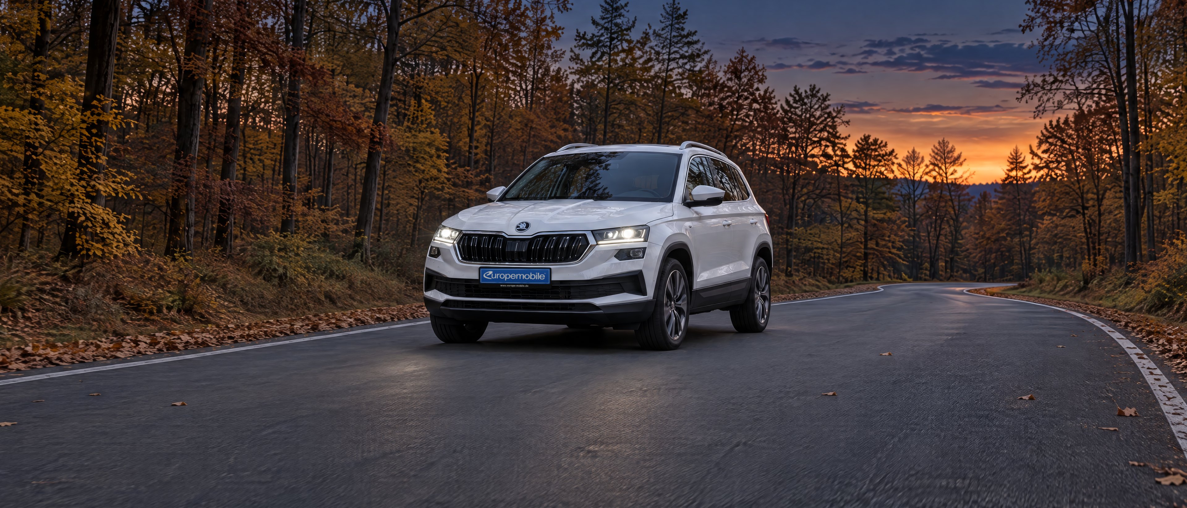 Weißer Škoda Karoq mit eingeschaltetem Abblendlicht auf einer Landstraße durch einen Herbstwald in der Abenddämmerung
