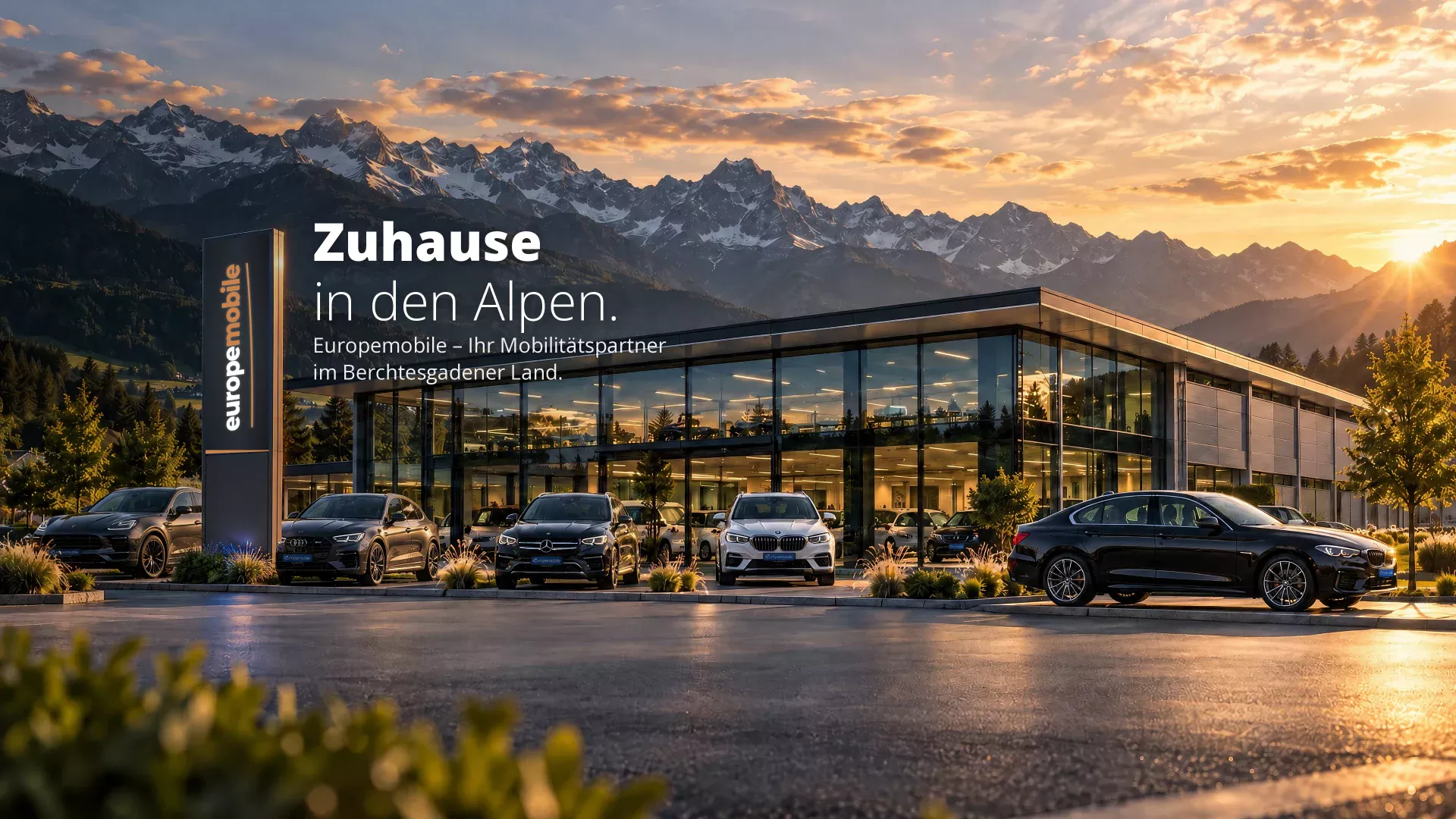 Zuhause in den Alpen: Europemobile-Autohaus mit Pylon und Fahrzeugen vor Bergpanorama bei Sonnenuntergang